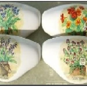 May include: Four white, oval-shaped decorative ceramic pieces. Each features a different floral design: blue forget-me-nots, orange and yellow primroses, purple lavender, and mixed flowers. The floral images are set against a yellow background.