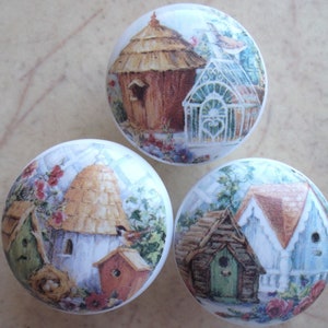 May include: Three round ceramic knobs with a white background and colorful images of birdhouses with thatched roofs, flowers, and greenery.