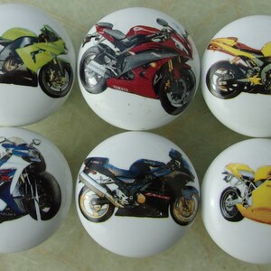 May include: Six ceramic cabinet knobs with images of different colored motorcycles. The motorcycles are all in a side profile view. The knobs are white with a glossy finish.