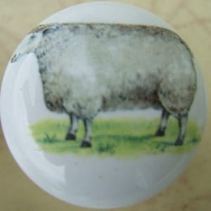 May include: White ceramic cabinet knob featuring a detailed illustration of a sheep standing on green grass. The sheep is depicted in shades of gray and brown, with a fluffy coat. The knob is round and has a glossy finish.