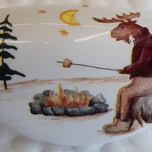 May include: A white, oval-shaped decorative item featuring a moose roasting a marshmallow over a campfire. The scene includes pine trees, a crescent moon, and stars, creating a cozy, outdoor theme.