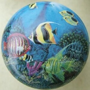 May include: A round decorative object featuring an underwater scene with colorful fish. The design includes a variety of fish in shades of yellow, black, red, and green, set against a blue background with coral and other aquatic elements.