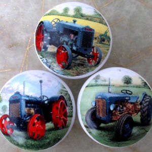 May include: Three ceramic knobs featuring vintage blue tractors with red wheels against a green field background. The knobs are approximately 2 inches in diameter.