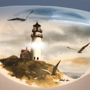 May include: A white, oval-shaped ceramic dish with a painted coastal scene. The artwork depicts a lighthouse on a hill, seagulls in flight, and a small house. The colors include white, blue, brown, and gray.