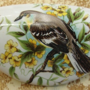 May include: A white ceramic knob with a painted design of a brown and white bird perched on a branch with yellow flowers.