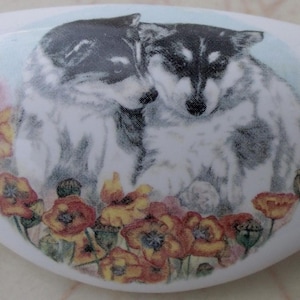 May include: White ceramic drawer pull featuring a watercolor illustration of two husky dogs nestled together. The dogs are surrounded by red, yellow, and pink poppies. The pull has a curved, elongated shape.