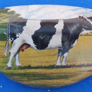 May include: Oval-shaped ceramic decorative piece featuring a black and white cow in a field. The cow is the focal point, set against a backdrop of a green field and trees under a cloudy sky. The edges are white.