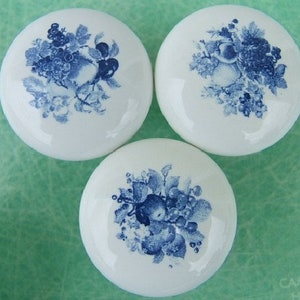 May include: Three white ceramic cabinet knobs with a glossy finish. Each knob features a blue floral and fruit design. The design includes apples, grapes, and leaves. The knobs are round and have a classic, vintage aesthetic.