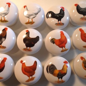 May include: A collection of white ceramic cabinet knobs, each featuring a different illustration of a rooster or hen. The birds are depicted in various colors, including black, brown, red, and white. These knobs are likely for kitchen or farmhouse decor.