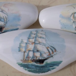May include: Three white, oval-shaped decorative objects with a nautical theme. Each features a detailed watercolor painting of a tall sailing ship on a blue ocean. The ships have white sails and dark hulls.