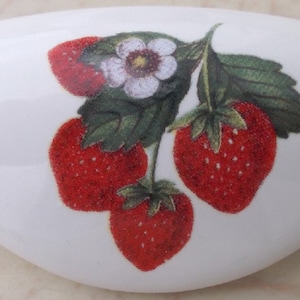 May include: White ceramic cabinet pull with a strawberry design. The strawberries are red with green leaves and a white flower. The pull is oval-shaped and has a glossy finish.
