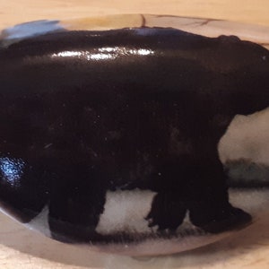 May include: Oval-shaped cabinet knob with a glossy white finish and a black bear silhouette. The bear is set against a backdrop of muted landscape colors. A decorative accent for furniture, drawers, or cabinets.
