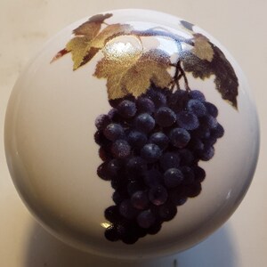 May include: A white, spherical decorative knob featuring a cluster of dark purple grapes and golden-hued leaves. The grapes are detailed with a glossy finish, and the leaves have a metallic sheen. This knob is likely for furniture or decor.