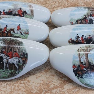 May include: Six white oval cabinet knobs with a hunting scene illustration. The knobs feature a glossy finish and a detailed image of people on horseback in a forest setting, with riders in red coats.