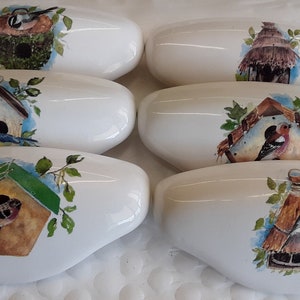 May include: Seven white, oval-shaped decorative stones with birdhouse and bird illustrations. Each stone features a different birdhouse design, with birds and foliage in watercolor style. The stones are arranged on a white, textured surface.