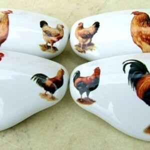 May include: Set of four white, oval-shaped cabinet knobs. Each knob features a detailed illustration of a rooster or hen in shades of brown, red, and black. The knobs are designed for furniture and home decor.