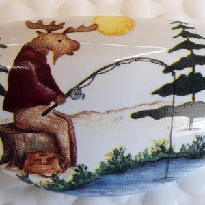 May include: White ceramic decorative piece with a painted scene of a moose fishing. The moose is wearing a maroon shirt and sitting on a tree stump. The background includes a yellow sun, pine trees, and a lake.