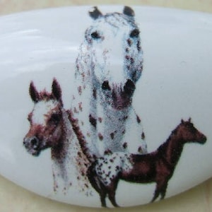 May include: A white ceramic oval-shaped ornament with a painted image of three spotted horses. The horses are brown, white, and black with brown spots.