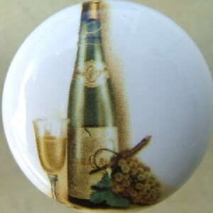 May include: White ceramic cabinet knob with a painted design featuring a wine bottle, a glass of wine, and a bunch of grapes. The bottle is green and gold, the wine is yellow, and the grapes are brown.