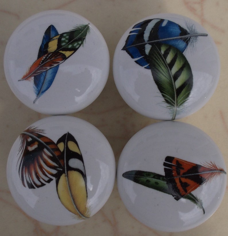 May include: Four white ceramic cabinet knobs with colourful feather designs. Each knob features a different arrangement of feathers in shades of blue, green, yellow, orange, and red. The knobs are round and have a glossy finish.