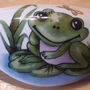 May include: White ceramic drawer pull featuring a cartoon frog sitting on a lily pad. The frog is green with a pink belly and is smiling. A yellow butterfly is above the frog's head, and green reeds are behind the frog.