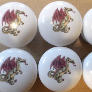 May include: Six white ceramic drawer knobs with a green dragon design. The dragons have red wings and are in a standing position with their tails curled.