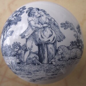 May include: A white ceramic knob with a blue and white design of a couple in a pastoral setting. The couple is standing in front of a tree and a sheep. The design is reminiscent of a vintage illustration.