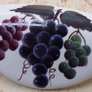 May include: White, oval-shaped decorative object with a painted design of three bunches of grapes. The grapes are purple, dark blue, and green, with green leaves. The object has a glossy finish.