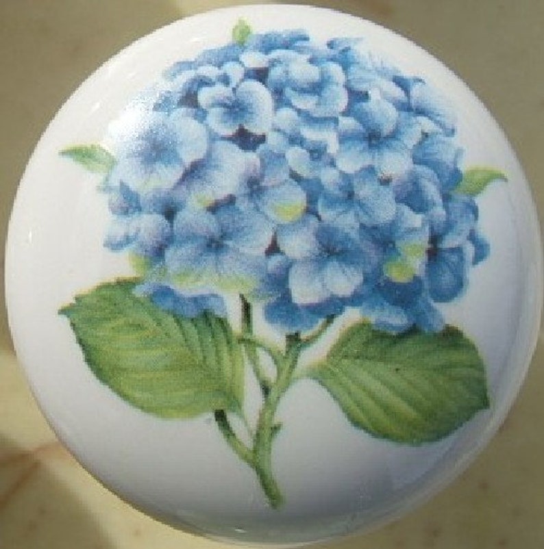 Op de afbeelding: Een witte keramische kastknop met een blauw hortensia bloemmotief. De bloemenafbeelding bevat een cluster van blauwe bloemblaadjes, groene bladeren en een groene stengel. De knop is rond en heeft een glanzende afwerking.
