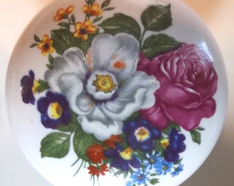 May include: A white ceramic knob with a floral design. The design features a pink rose, a white flower, and purple and yellow flowers. The background is white.