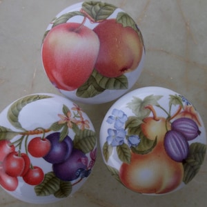 May include: Three ceramic knobs with painted fruit designs. The first knob features a red apple and a yellow pear with green leaves. The second knob features a purple plum, red cherries, and green leaves. The third knob features a yellow pear, purple figs, and blue flowers.