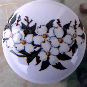 May include: White ceramic knob with a floral design featuring white dogwood blossoms with yellow centers and green leaves.