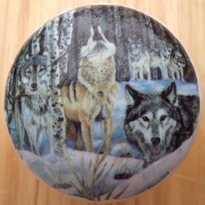May include: Round decorative knob featuring a detailed image of a wolf pack in a snowy forest. The wolves are depicted in shades of gray, brown, and white, set against a backdrop of birch trees and a winter landscape. The knob is white.