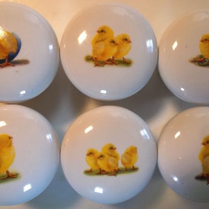May include: Six white ceramic knobs with yellow chick designs. Each knob features a different chick image, including a chick hatching from an egg, a group of chicks, and a single chick.