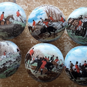 May include: Six round decorative objects with hunting scenes. Each object features a different image of riders on horseback in red coats, with trees and a blue sky background. The objects are arranged on a light brown surface.