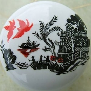 May include: A white ceramic knob with a black and red silhouette design of a Chinese landscape. The design features a pagoda, a bridge, a boat, trees, and birds.