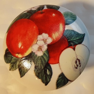 May include: A white ceramic knob with a red apple design. The knob features three red apples with green leaves and white flowers.