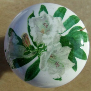 May include: A white ceramic cabinet knob with a floral design. The knob features a detailed print of white flowers with green leaves and stems. The knob is round and has a glossy finish.