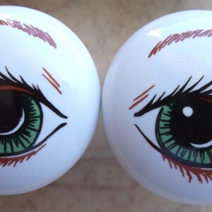 May include: Pair of white ceramic drawer knobs with a green eye design. Each knob features a black pupil, brown eyelashes, and a red eyebrow.