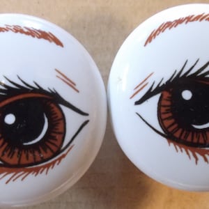 May include: Two white ceramic knobs with brown painted eyes. Each knob features a brown eye with a black pupil and a black eyelash.