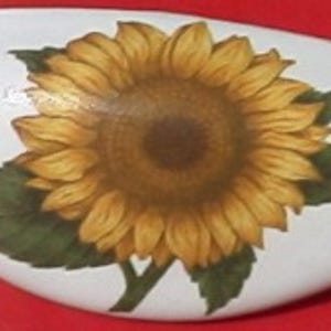 May include: White, oblong decorative stone featuring a detailed sunflower design. The sunflower has yellow petals, a brown center, green leaves, and a green stem. The stone is smooth and appears to be painted.
