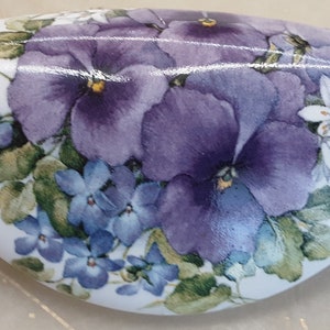 May include: A white ceramic heart-shaped dish with a painted design of purple and blue pansies and white flowers. The dish is decorated with green leaves and has a glossy finish.