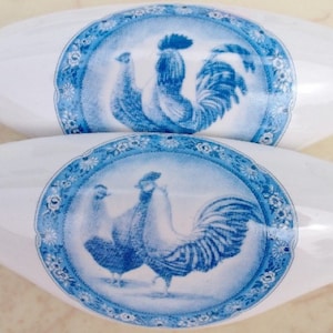 May include: Two white ceramic cabinet knobs with a blue and white rooster and hen design. The oval knobs have a blue floral border around the central image. These decorative knobs are ideal for adding a touch of farmhouse style to furniture.