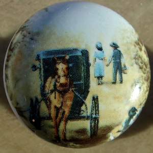 May include: A round, off-white cabinet knob with a painted scene. The image depicts a horse-drawn carriage, a couple holding hands, and a person in the background. The knob has a glossy finish.