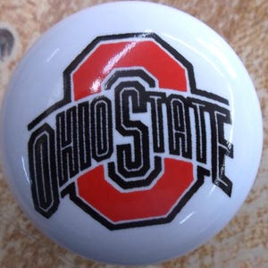 May include: A round, white ceramic knob featuring the Ohio State University logo printed in the center. The design includes a large red block letter O with the word OHIO STATE written in black, bold, stylized lettering across the front surface.