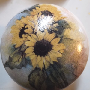 May include: A round ceramic knob with a watercolor design of sunflowers in shades of yellow, brown, and green on a white background.