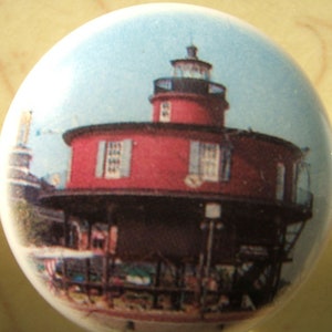 May include: A round, light blue button featuring a red lighthouse with a black roof and a white tower. The lighthouse has two windows with blue shutters. The button has a glossy finish.