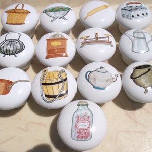 May include: A collection of white ceramic cabinet knobs, each featuring a different hand-painted kitchen-themed design. Designs include a basket, colander, rolling pin, iron, scale, teapot, and a jar filled with hearts. These knobs add a charming, vintage touch.