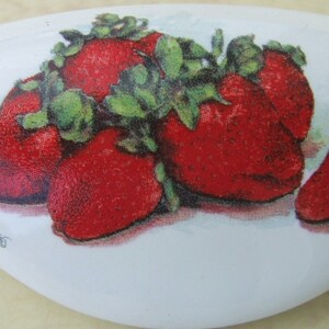 May include: A white ceramic knob with a red and green strawberry design. The knob is shaped like a teardrop and has a smooth, glossy finish.