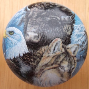May include: A round wooden ornament with a colorful image of a bald eagle, a wolf, and a bison. The background is a blue sky with clouds.